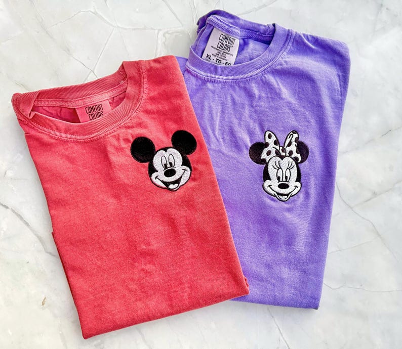May include: Two Comfort Colors t-shirts, one coral and one purple, each with a cartoon character face embroidered on the chest. The coral shirt features Mickey Mouse, and the purple shirt features Minnie Mouse.