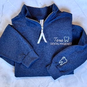 May include: A navy blue quarter-zip sweatshirt with white embroidered text and a tooth graphic. The text reads "Tara Dental Hygienist." A tooth graphic is also on the sleeve. The zipper has a white pull.