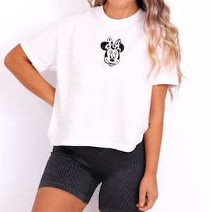 Peut inclure: T-shirt blanc avec un motif brod&eacute; de Minnie Mouse en noir et blanc, avec un n&oelig;ud &agrave; pois. Le t-shirt est port&eacute; avec un short ray&eacute; noir et gris. L'ensemble a un style d&eacute;contract&eacute; et confortable.