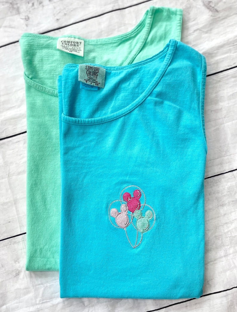 May include: Two sleeveless t-shirts, one mint green and one turquoise, are displayed. The turquoise shirt features an embroidered design of Mickey Mouse balloons. Both shirts have the "Comfort Colors" label.