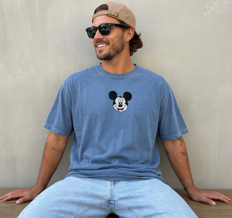 May include: A blue short-sleeved t-shirt featuring a small, centered black and white graphic of the Mickey Mouse character face on the chest. The shirt has a crew neckline and a relaxed fit, constructed from a solid-colored cotton fabric.