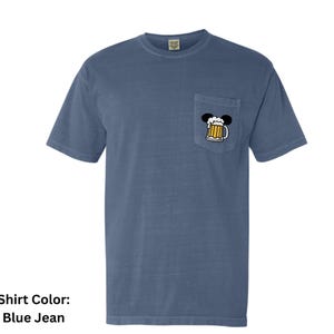 May include: A blue jean colored short-sleeve t-shirt with a crew neckline and a single chest pocket. The pocket features a graphic of a beer mug with white foam, topped with two black circular ears, creating a silhouette of a mouse head.