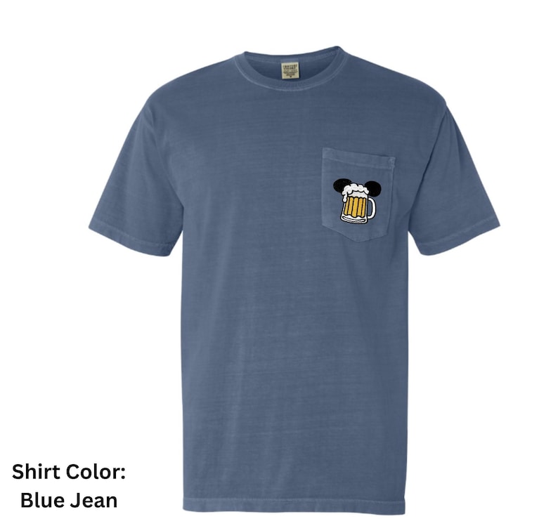 May include: A blue jean colored short-sleeve t-shirt with a crew neckline and a single chest pocket. The pocket features a graphic of a beer mug with white foam, topped with two black circular ears, creating a silhouette of a mouse head.