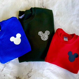 May include: Three sweatshirts with different colored Mickey Mouse designs. The first sweatshirt is blue with a white Mickey Mouse. The second sweatshirt is green with a white Mickey Mouse with a leopard print. The third sweatshirt is red with a black Mickey Mouse.