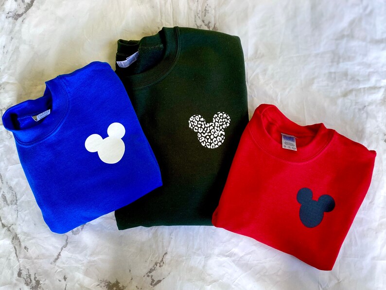 May include: Three sweatshirts with different colored Mickey Mouse designs. The first sweatshirt is blue with a white Mickey Mouse. The second sweatshirt is green with a white Mickey Mouse with a leopard print. The third sweatshirt is red with a black Mickey Mouse.