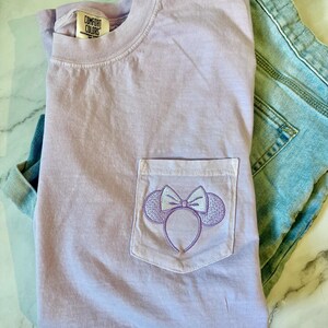 Op de afbeelding: Een lavendelkleurig T-shirt met een zakje met een geborduurd Minnie Mouse-oorband. Het T-shirt heeft de tekst "Comfort Colors" op het label.
