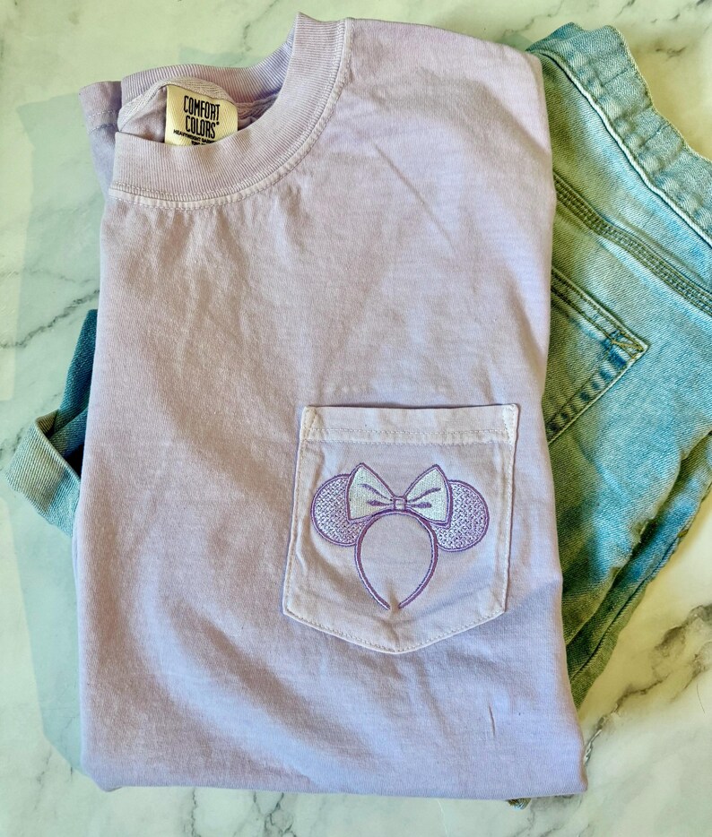 Op de afbeelding: Een lavendelkleurig T-shirt met een zakje met een geborduurd Minnie Mouse-oorband. Het T-shirt heeft de tekst "Comfort Colors" op het label.