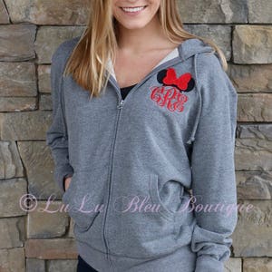 Puede incluir: Una sudadera gris con cremallera y capucha con un lazo rojo y un monograma en la parte delantera. La sudadera la lleva puesta una persona de pie frente a una pared de piedra.