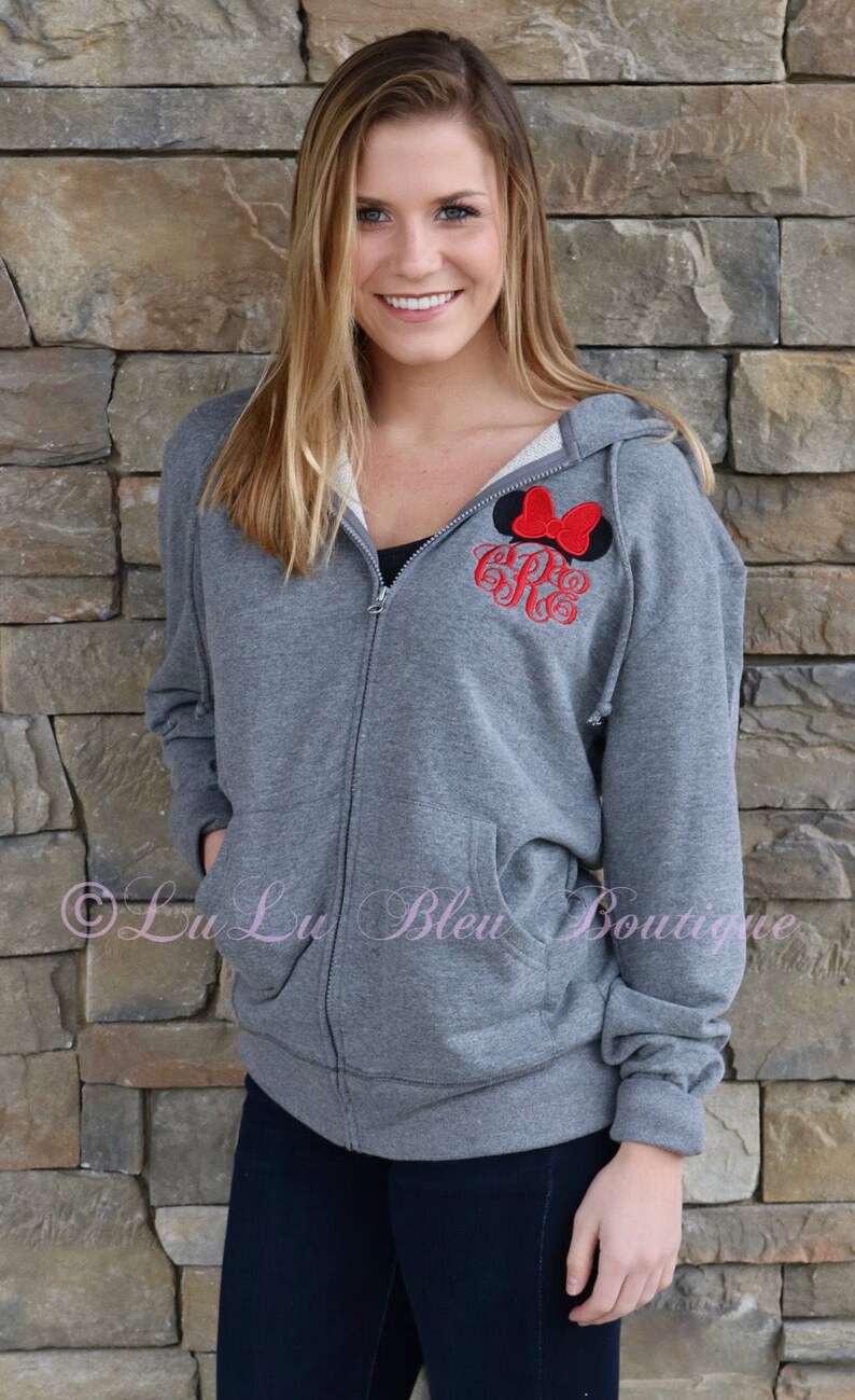 Puede incluir: Una sudadera gris con cremallera y capucha con un lazo rojo y un monograma en la parte delantera. La sudadera la lleva puesta una persona de pie frente a una pared de piedra.