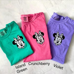 May include: Three Comfort Colors t-shirts in Island Green, Crunchberry, and Violet. Each shirt features a Minnie Mouse graphic with a black and white polka dot bow. The shirt colors are labeled with text.
