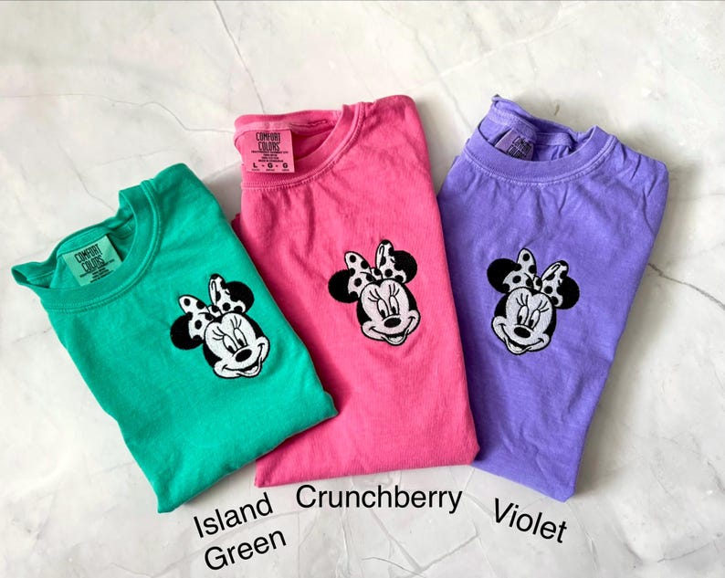 May include: Three Comfort Colors t-shirts in Island Green, Crunchberry, and Violet. Each shirt features a Minnie Mouse graphic with a black and white polka dot bow. The shirt colors are labeled with text.