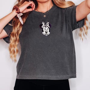 Peut inclure: T-shirt gris fonc&eacute; avec une broderie de Minnie Mouse sur la poitrine. Le motif comprend un visage blanc, des yeux noirs et un n&oelig;ud violet &agrave; pois blancs. Le t-shirt a des manches courtes et un aspect l&eacute;g&egrave;rement d&eacute;lav&eacute;.
