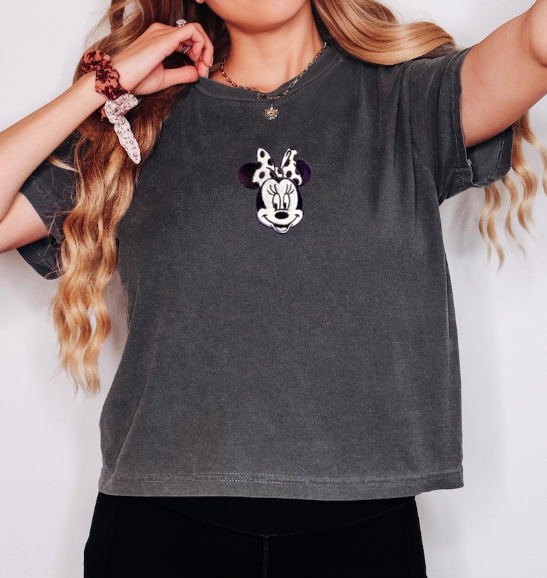 Peut inclure: T-shirt gris fonc&eacute; avec une broderie de Minnie Mouse sur la poitrine. Le motif comprend un visage blanc, des yeux noirs et un n&oelig;ud violet &agrave; pois blancs. Le t-shirt a des manches courtes et un aspect l&eacute;g&egrave;rement d&eacute;lav&eacute;.