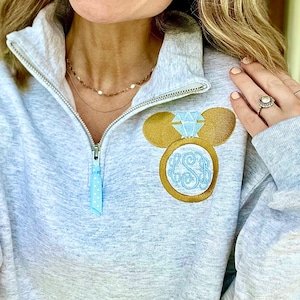 Pu&ograve; includere: Una felpa grigia con un design a forma di testa di Topolino dorata con un anello di diamanti blu. L'anello ha le iniziali "LSB" all'interno.