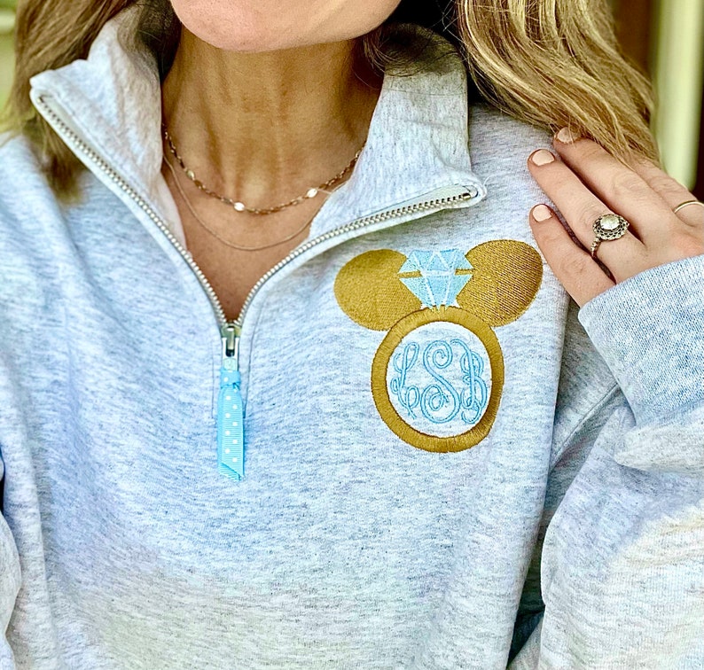 Pu&ograve; includere: Una felpa grigia con un design a forma di testa di Topolino dorata con un anello di diamanti blu. L'anello ha le iniziali "LSB" all'interno.