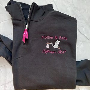 Puede incluir: Sudadera negra con cremallera de un cuarto de cuello con dise&ntilde;o de cig&uuml;e&ntilde;a bordada que lleva un beb&eacute;. Los textos "Mother & Baby" y "Tiffany, RN" est&aacute;n bordados en rosa. Una cinta rosa con lunares est&aacute; unida a la cremallera.