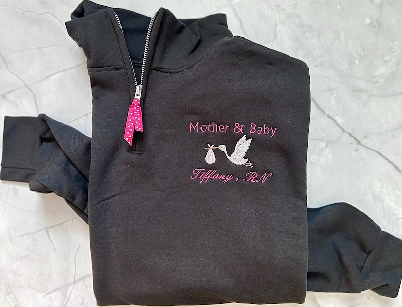 Puede incluir: Sudadera negra con cremallera de un cuarto de cuello con dise&ntilde;o de cig&uuml;e&ntilde;a bordada que lleva un beb&eacute;. Los textos "Mother & Baby" y "Tiffany, RN" est&aacute;n bordados en rosa. Una cinta rosa con lunares est&aacute; unida a la cremallera.