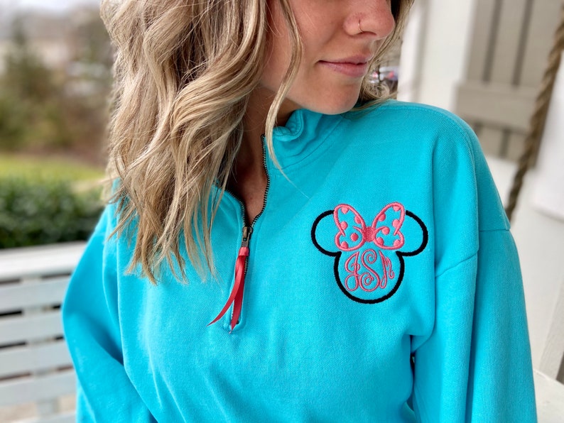 May include: A turquoise blue sweatshirt with a black and pink embroidered Minnie Mouse head with a bow. The sweatshirt has a red zipper.
