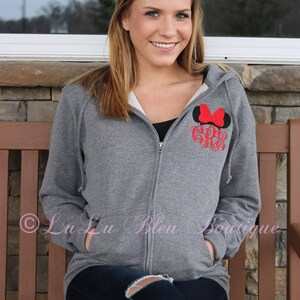 Puede incluir: Una sudadera gris con cremallera con un dise&ntilde;o bordado de orejas de Mickey Mouse en negro y rojo y un monograma. La sudadera tiene capucha y dos bolsillos.