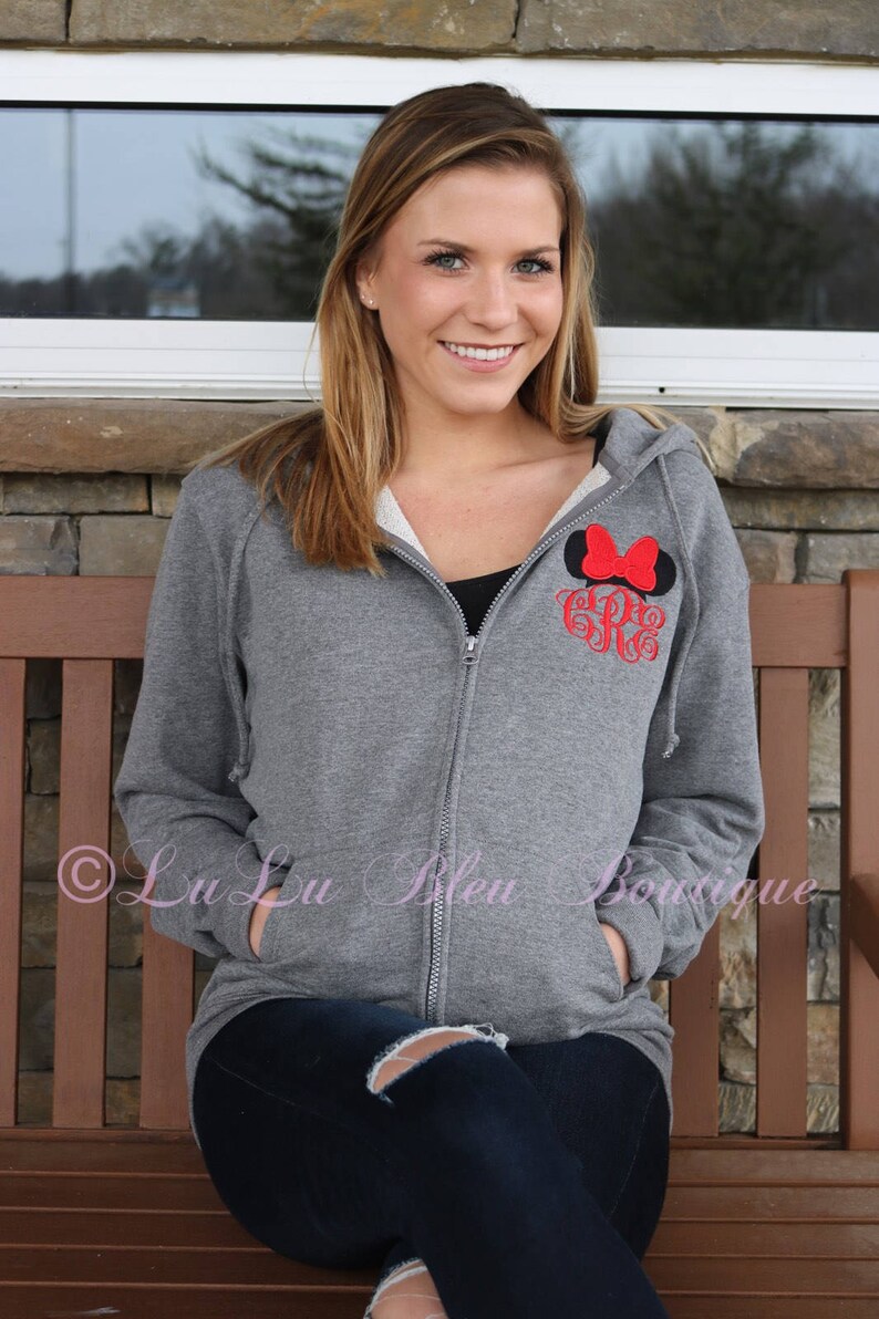 Puede incluir: Una sudadera gris con cremallera con un dise&ntilde;o bordado de orejas de Mickey Mouse en negro y rojo y un monograma. La sudadera tiene capucha y dos bolsillos.