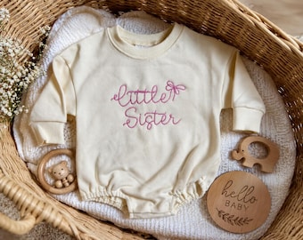 Puede incluir: Un pelele de bebé de color crema con mangas largas y el texto bordado en rosa "little Sister" con un pequeño lazo. La prenda tiene puños acanalados y cuello redondo, presentada junto a un juguete de madera con forma de elefante.
