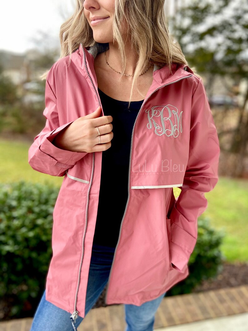 May include: A pink hooded raincoat with a white zipper and reflective trim. The raincoat has a monogrammed "KRM" on the front. The text "Lulu Bleu" is visible on the bottom right of the raincoat.