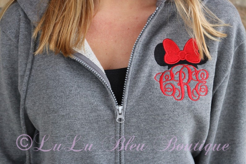 Puede incluir: Sudadera gris con cremallera con una cabeza de Minnie Mouse bordada en rojo y negro y unas iniciales bordadas. La sudadera tiene un forro de polar blanco.