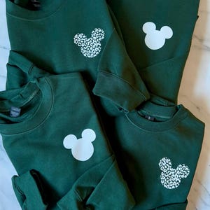 May include: Four green sweatshirts with white Mickey Mouse designs. One sweatshirt has a solid white Mickey Mouse, one has a leopard print Mickey Mouse, and two have a solid white Mickey Mouse.