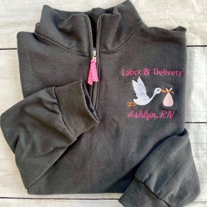 Puede incluir: Sudadera gris oscuro con cremallera de cuarto de cremallera con texto bordado y gr&aacute;fico de cig&uuml;e&ntilde;a. El texto dice "Labor & Delivery" encima de una cig&uuml;e&ntilde;a que lleva un paquete de beb&eacute;, con el nombre "Ashlyn, RN" debajo. El tirador de la cremallera tiene una cinta rosa de lunares.