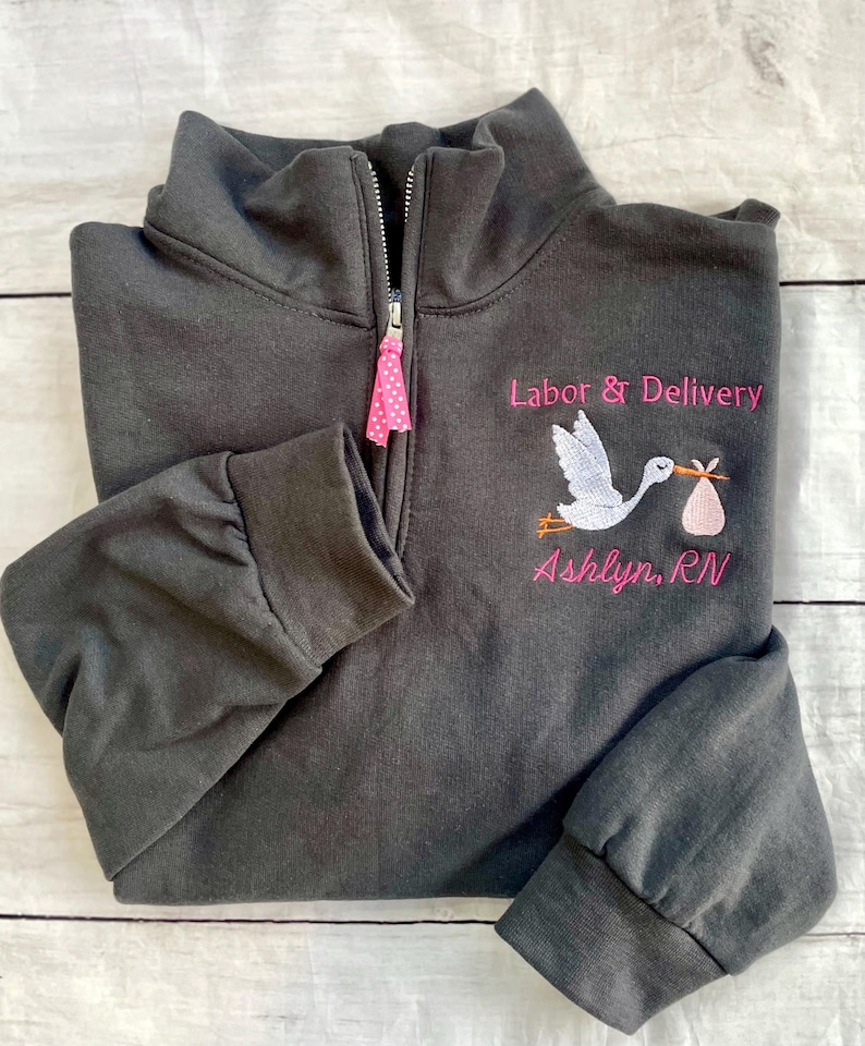 Puede incluir: Sudadera gris oscuro con cremallera de cuarto de cremallera con texto bordado y gr&aacute;fico de cig&uuml;e&ntilde;a. El texto dice "Labor & Delivery" encima de una cig&uuml;e&ntilde;a que lleva un paquete de beb&eacute;, con el nombre "Ashlyn, RN" debajo. El tirador de la cremallera tiene una cinta rosa de lunares.