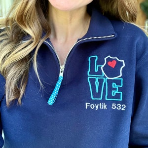 Peut inclure: Un sweat-shirt bleu marine à fermeture éclair quart avec un motif brodé turquoise qui dit "LOVE" avec un cœur rouge à l'intérieur du "O". Le texte "Foytik 532" est brodé sous le motif.
