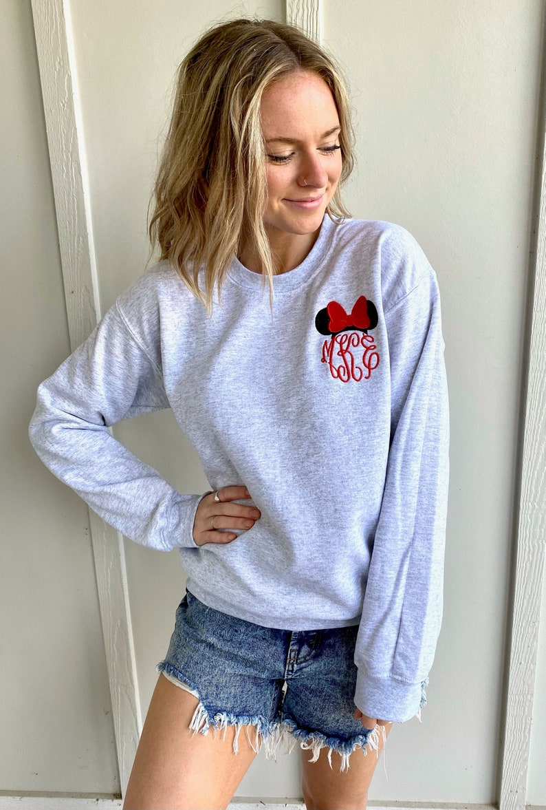 Puede incluir: Una sudadera gris con orejas de Mickey Mouse y un lazo bordados en rojo y negro en el pecho izquierdo. La sudadera tiene cuello redondo y mangas largas.