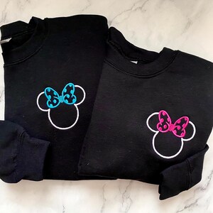 May include: Two black sweatshirts with white outlines of Minnie Mouse's head. One sweatshirt has a blue polka dot bow and the other has a pink polka dot bow.