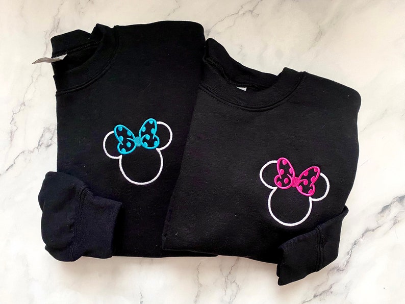 May include: Two black sweatshirts with white outlines of Minnie Mouse's head. One sweatshirt has a blue polka dot bow and the other has a pink polka dot bow.
