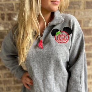 Op de afbeelding: Een grijs sweatshirt met een rood en groen appelontwerp met een monogram op de appel. De appel wordt omringd door zwarte Mickey Mouse-oren.