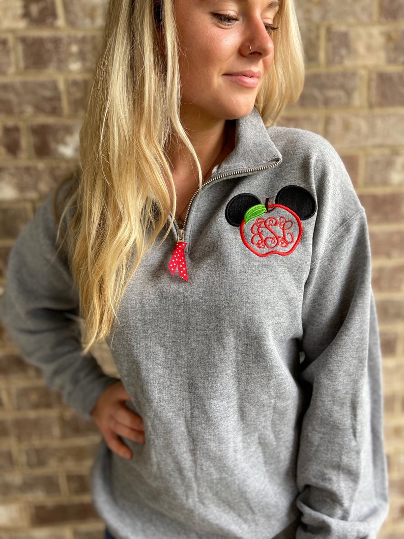 Op de afbeelding: Een grijs sweatshirt met een rood en groen appelontwerp met een monogram op de appel. De appel wordt omringd door zwarte Mickey Mouse-oren.