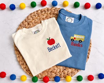 Pode incluir: Duas camisetas de manga curta e gola redonda, uma na cor creme e outra azul. A camiseta creme apresenta uma maçã vermelha bordada e o nome Beckett em azul. A camiseta azul apresenta um ônibus escolar amarelo bordado e o nome Banks em vermelho.