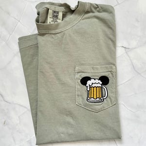 May include: A sage green short-sleeve t-shirt with a chest pocket featuring an embroidered graphic of a beer mug with white foam and black mouse ears. The inner neck label displays the text Comfort Colors, Heavyweight Garment Dye, 100% Cotton.