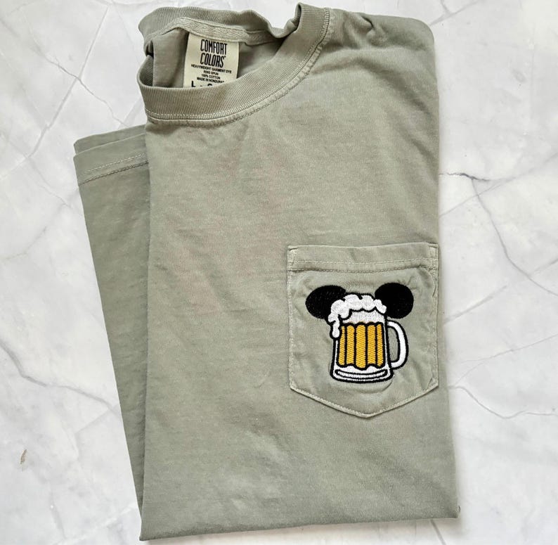 May include: A sage green short-sleeve t-shirt with a chest pocket featuring an embroidered graphic of a beer mug with white foam and black mouse ears. The inner neck label displays the text Comfort Colors, Heavyweight Garment Dye, 100% Cotton.