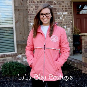 May include: A pink rain jacket with a white zipper and a white monogram on the left chest. The jacket has a hood and two pockets.