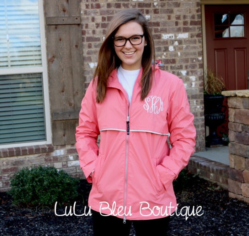 May include: A pink rain jacket with a white zipper and a white monogram on the left chest. The jacket has a hood and two pockets.