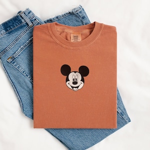 May include: A terracotta-colored short-sleeve t-shirt featuring a black and white embroidered Mickey Mouse head graphic centered on the chest. The shirt has a crew neckline and a Comfort Colors brand label printed on the inside of the collar.