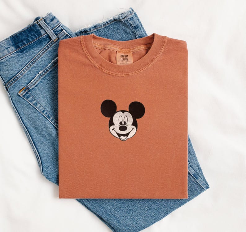 May include: A terracotta-colored short-sleeve t-shirt featuring a black and white embroidered Mickey Mouse head graphic centered on the chest. The shirt has a crew neckline and a Comfort Colors brand label printed on the inside of the collar.