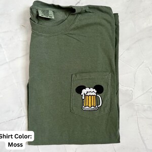 May include: A moss green short-sleeve t-shirt with a crew neckline and a single chest pocket. The pocket features an embroidered graphic of a beer mug with white foam, topped with two black circular ears. The shirt has a Comfort Colors label.