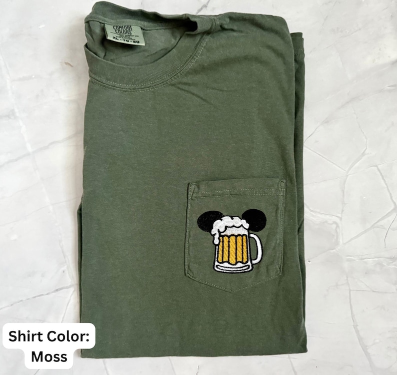 May include: A moss green short-sleeve t-shirt with a crew neckline and a single chest pocket. The pocket features an embroidered graphic of a beer mug with white foam, topped with two black circular ears. The shirt has a Comfort Colors label.
