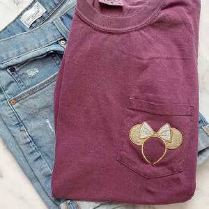 Op de afbeelding: Een maroon t-shirt met een geborduurd Minnie Mouse-orenontwerp in goud en wit op de zak.