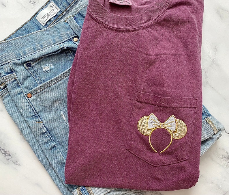 Op de afbeelding: Een maroon t-shirt met een geborduurd Minnie Mouse-orenontwerp in goud en wit op de zak.