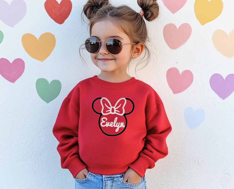 May include: A red sweatshirt featuring a black and white Mickey Mouse head design with the name "Evelyn" in white. The sweatshirt is being worn by a young girl with brown hair and sunglasses.