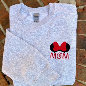 Puede incluir: Sudadera gris con un lazo rojo y orejas negras, y la palabra "MOM" bordada en la parte delantera.