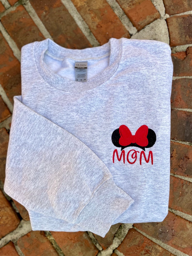 Puede incluir: Sudadera gris con un lazo rojo y orejas negras, y la palabra "MOM" bordada en la parte delantera.